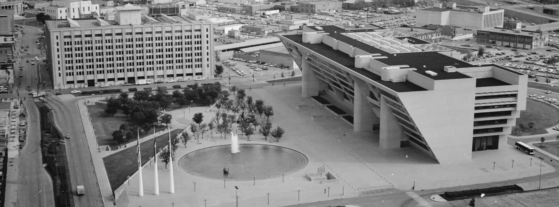 Aerial photograph looking east across City Hall Plaza. Dallas City Hall sits at right, its upper floors cantilevered out over the plaza; a circular fountain, three flagpoles, and rows of young trees fill the open ground in front of it.